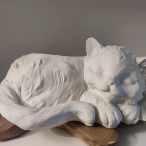 Op de afbeelding: Een witte kattenbeeldhouwwerk in een slaaphouding, rustend op een houten voet. De vacht van de kat is getextureerd en de ogen zijn gesloten. Het beeldhouwwerk is een decoratief item, waarschijnlijk voor de interieurdecoratie.