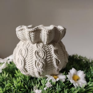 Peut inclure: Un vase crème avec un motif tricoté, orné d'un dessus froncé et de motifs en forme de cœur. Le vase est posé sur de l'herbe artificielle verte, décorée de marguerites blanches. Le vase a une surface texturée.