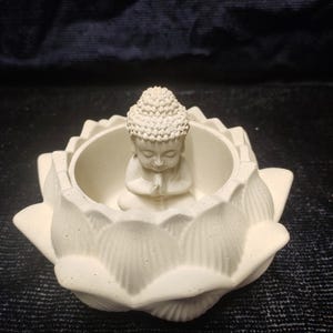 Op de afbeelding: Een wit Boeddha-beeld in een lotusbloemvormige container. De Boeddha zit in een zittende positie met de handen gevouwen in gebed. De lotusbloem heeft grote, getextureerde bloembladen. Het beeld is gemaakt van licht materiaal.