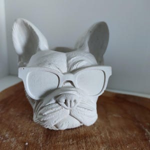 Puede incluir: Una maceta de hormigón gris claro con forma de bulldog francés con gafas de sol. La cara del perro está detallada con arrugas y una superficie texturizada. La maceta tiene una parte superior ahuecada para guardar plantas u otros objetos.