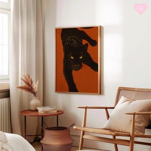 May include: A framed art print of a black panther against an orange backdrop. The panther is shown in a dynamic pose, with yellow eyes. The artwork is displayed in a room with neutral-toned furniture and decor.