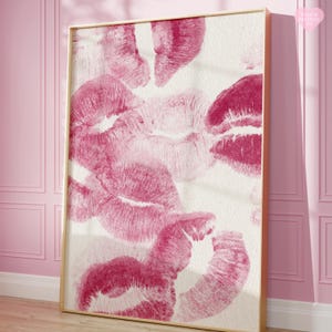 May include: A framed art print featuring multiple lip imprints in varying shades of pink and magenta against a white background. The artwork is displayed in a gold-colored frame, creating a romantic and artistic aesthetic.