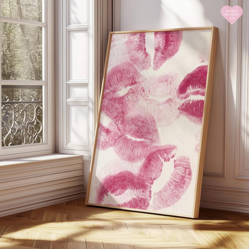 May include: A framed art print featuring multiple pink lipstick kiss marks on a white background. The artwork is displayed in a natural wood frame, leaning against a white wall near a window.