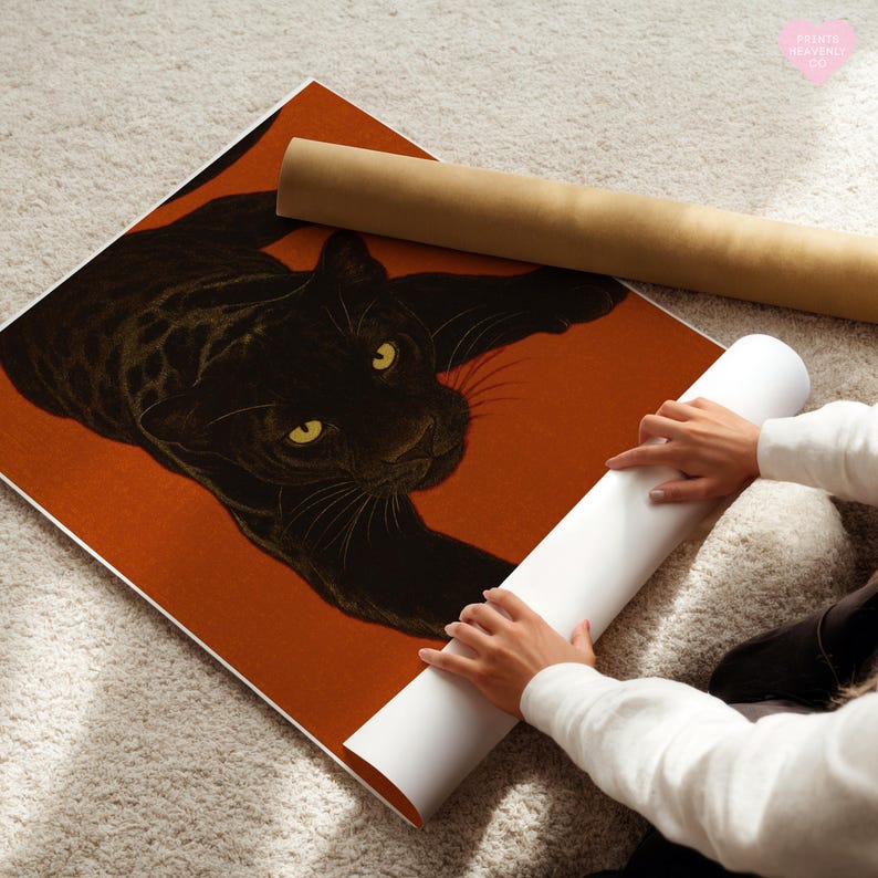 May include: A black panther art print with yellow eyes on an orange background. A rolled-up poster is next to the print. The print is on a white surface. The words "Painted Heavenly Co" are in the upper right corner.