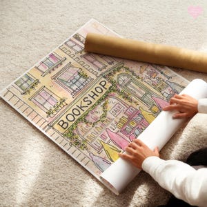 May include: Watercolor print of a bookstore with the word "BOOKSHOP" in large letters. The print features a detailed illustration of a building with windows, a door, and a "SALE" sign. A rolled-up poster and a person's hands are also visible.