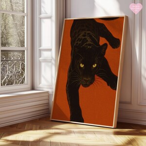 May include: A framed art print showing a black panther on an orange backdrop. The panther is illustrated in a descending pose, with yellow eyes. The artwork is displayed in a room with natural light, near a window.