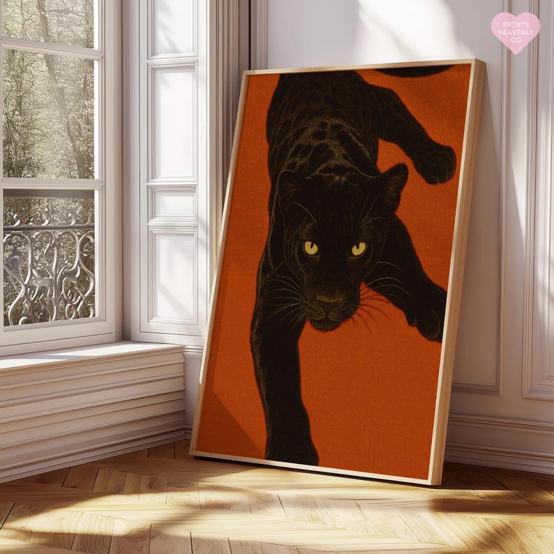 May include: A framed art print showing a black panther on an orange backdrop. The panther is illustrated in a descending pose, with yellow eyes. The artwork is displayed in a room with natural light, near a window.