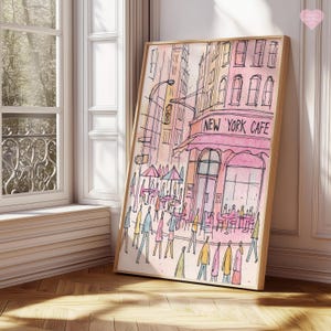 May include: Watercolor print of a New York Cafe scene. The artwork features a pink awning with the cafe's name, figures of people, and tables with umbrellas. The buildings are in shades of pink, yellow, and orange, framed in a light wood.