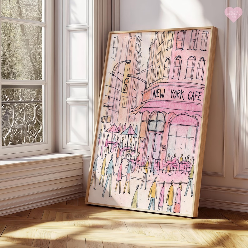 May include: Watercolor print of a New York Cafe scene. The artwork features a pink awning with the cafe's name, figures of people, and tables with umbrellas. The buildings are in shades of pink, yellow, and orange, framed in a light wood.