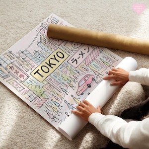 May include: A colorful illustration of Tokyo with the city name in a gold rectangle. The artwork includes Japanese text, a pink car, and figures of people. A rolled-up poster and a brown tube are also visible.