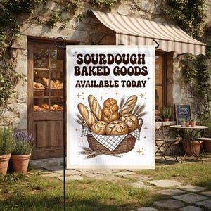 May include: A rectangular garden flag featuring the brown text SOURDOUGH BAKED GOODS AVAILABLE TODAY above an illustration of a woven basket filled with various loaves of bread, including baguettes and round boules, accented by small gold stars.