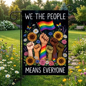 May include: A rectangular garden flag with a black background featuring the white text WE THE PEOPLE MEANS EVERYONE. The design displays five raised fists of varying skin tones, one holding a rainbow flag, surrounded by sunflowers and butterflies.