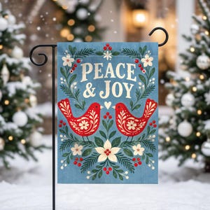 May include: A rectangular garden flag featuring a blue background with the text PEACE & JOY in gold lettering. Two red birds with white floral patterns face each other, surrounded by green pine branches, white flowers, and red berries.