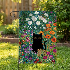May include: A garden flag featuring a black cat with yellow eyes, surrounded by colorful flowers. The word "Welcome" is written in white. The flag is displayed on a black metal stand, set against a backdrop of green grass and foliage.