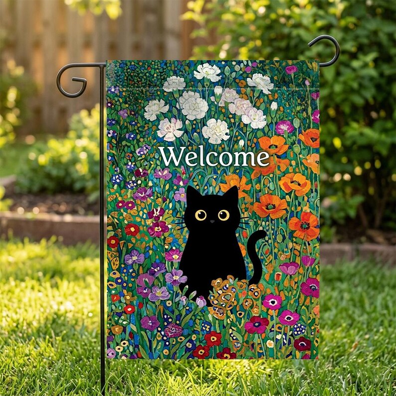 May include: A garden flag featuring a black cat with yellow eyes, surrounded by colorful flowers. The word "Welcome" is written in white. The flag is displayed on a black metal stand, set against a backdrop of green grass and foliage.