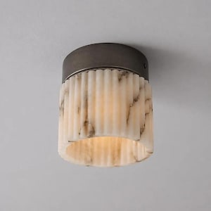 May include: A flush mount ceiling light fixture featuring a cylindrical, fluted shade made of white and grey marbled stone. The shade is capped with a dark bronze-toned metal base that secures the light to the ceiling surface.