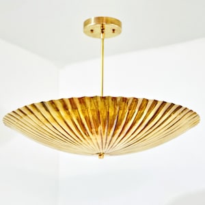 May include: A gold-toned pendant light fixture featuring a wide, circular, pleated shade with a metallic finish. The fixture is suspended from a thin, vertical rod attached to a matching circular ceiling canopy, creating a sleek, uniform appearance.