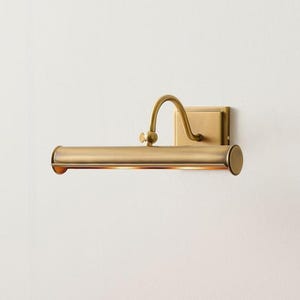 May include: A wall-mounted picture light with a brushed brass finish. The fixture features a rectangular backplate, a curved adjustable arm, and a horizontal cylindrical lamp head with an open bottom that emits a warm glow of light.