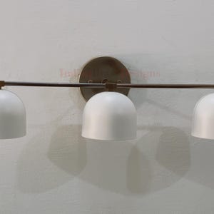 Puede incluir: Una moderna lámpara de tocador de tres luces. Cuenta con tres pantallas blancas en forma de cúpula unidas a una barra horizontal. La barra y la placa de montaje tienen un acabado de latón. La lámpara está montada en una pared texturizada de color blanquecino.