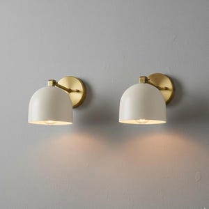 May include: Two wall sconces featuring cream-coloured, dome-shaped shades and brass-toned arms and backplates. The sconces are lit, emitting a warm light. The design is minimalist and modern, suitable for various interior styles.
