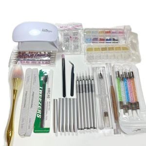 K&ouml;nnte beinhalten: Ein Set mit Nageldesign-Werkzeugen und Zubeh&ouml;r, darunter eine wei&szlig;e SUNmini UV-Lampe, Nagelfeilen, Dotting-Tools, Pinzetten und verschiedene Verzierungen. Das Set enth&auml;lt einen Pinsel mit goldenem Griff und eine Auswahl an Nageldesign-Zubeh&ouml;r.