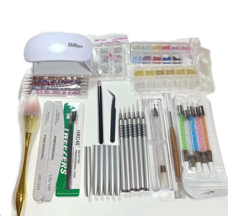 K&ouml;nnte beinhalten: Ein Set mit Nageldesign-Werkzeugen und Zubeh&ouml;r, darunter eine wei&szlig;e SUNmini UV-Lampe, Nagelfeilen, Dotting-Tools, Pinzetten und verschiedene Verzierungen. Das Set enth&auml;lt einen Pinsel mit goldenem Griff und eine Auswahl an Nageldesign-Zubeh&ouml;r.