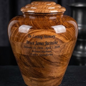 May include: A wooden urn with a glossy finish, featuring a rounded shape and a matching lid. The urn has a natural wood grain pattern and includes engraved text: "In Loving Memory Bruce James Jacintho" with dates and titles. A dignified memorial.
