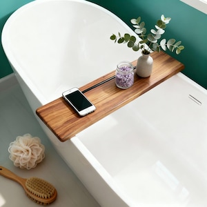 Peut inclure: Un plateau de bain en bois avec une barre métallique noire, contenant un smartphone, un petit vase avec de la verdure et un pot de sels de bain violets. Une baignoire blanche est au premier plan, avec un luffa et une brosse en bois.