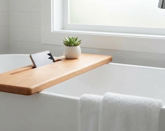 Peut inclure: Une baignoire blanche avec un plateau de bain en bois contenant un téléphone et une petite plante succulente en pot. Deux serviettes blanches sont accrochées sur le côté de la baignoire. Un robinet chromé et une fenêtre sont en arrière-plan.