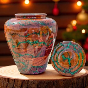 May include: A handcrafted wooden urn and lid with a unique design. The urn is a rich brown with vibrant green accents, and the lid mirrors the design. The urn and lid are displayed on a wooden slice, with a blurred background of Christmas lights.