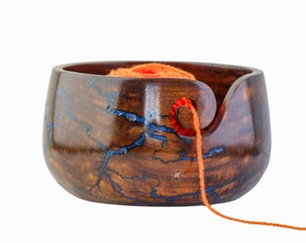 Könnte beinhalten: Handgefertigte Garnschale aus Holz mit brauner Oberfläche und blauen Harzakzenten. Die Schale enthält einen orangefarbenen Garnknäuel, wobei der Faden durch einen gebogenen Schlitz austritt. Ein praktisches Accessoire für Stricker und Häkler.