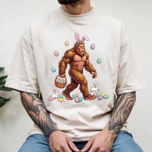 May include: Beige t-shirt featuring a cartoon Bigfoot wearing bunny ears and carrying a basket of Easter eggs. The design includes colorful Easter eggs and running rabbits, creating a festive and whimsical theme.