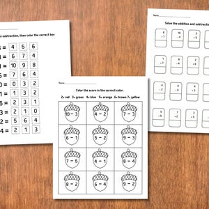 May include: Three worksheets with math problems for children. One sheet has subtraction problems, another has addition and subtraction problems, and the third has acorn coloring by number. The worksheets are on a wooden surface.