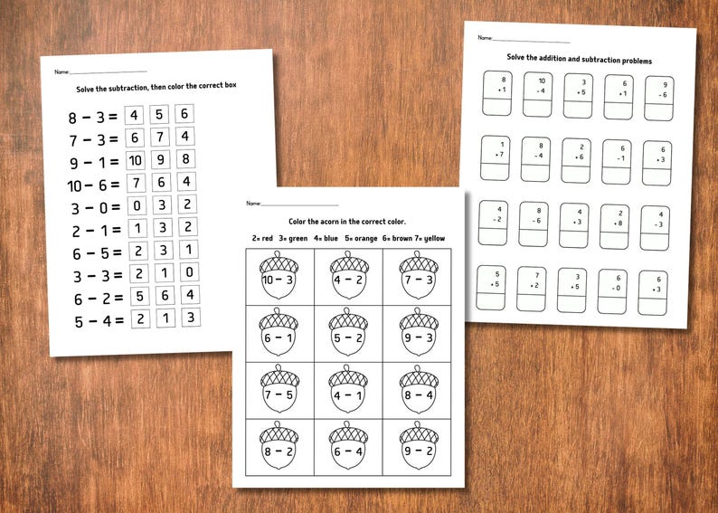 May include: Three worksheets with math problems for children. One sheet has subtraction problems, another has addition and subtraction problems, and the third has acorn coloring by number. The worksheets are on a wooden surface.