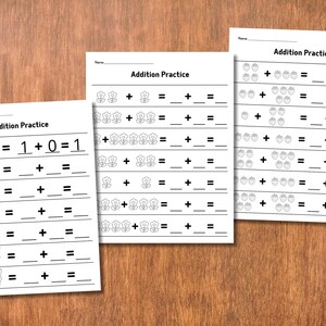 May include: Three white sheets of paper with "Addition Practice" printed on them. The sheets feature addition problems with hand gestures, flowers, and strawberries as visual aids. The background is a wooden surface.