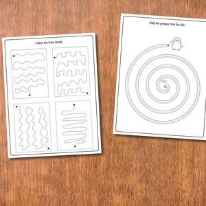 May include: Two white activity sheets with black line drawings on a wooden surface. One sheet has line tracing exercises and the text "Follow the lines slowly." The other sheet has a spiral maze with a penguin and the text "Help the penguin find the fish."