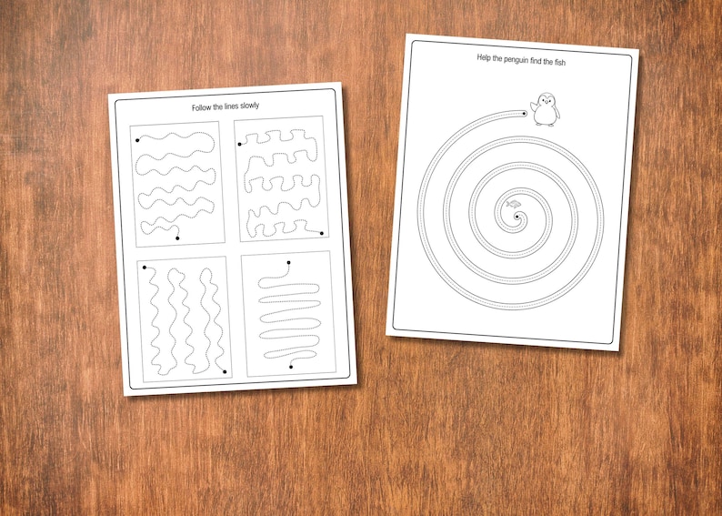 May include: Two white activity sheets with black line drawings on a wooden surface. One sheet has line tracing exercises and the text "Follow the lines slowly." The other sheet has a spiral maze with a penguin and the text "Help the penguin find the fish."
