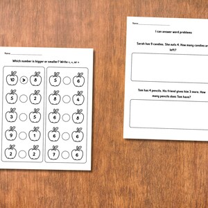 May include: Two white worksheets on a wooden surface. One worksheet asks "Which number is bigger or smaller?" with apple illustrations and number comparisons. The other asks word problems about candies and pencils.