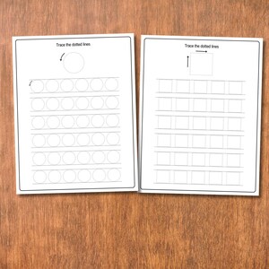 May include: Two white worksheets with black line drawings and text on a wooden surface. The worksheets have dotted lines for tracing shapes and the text "Trace the dotted lines". One worksheet has circles, the other has squares.