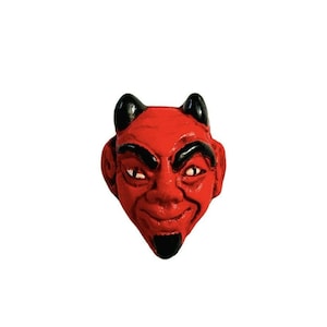 May include: A red devil face with black horns, eyebrows, and goatee. The face has a mischievous expression with white eyes and a slight smile. The devil face is a decorative item.