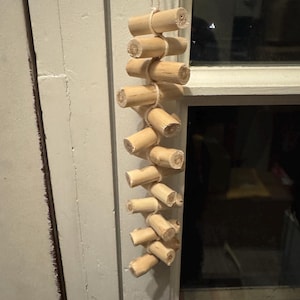 Puede incluir: Un juguete colgante para animales pequeños que consiste en una serie de troncos de madera cilíndricos de color claro conectados por un cordel grueso de color blanco roto. Los troncos están dispuestos en un patrón vertical escalonado.