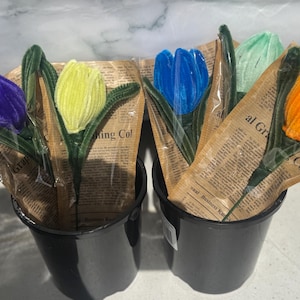 May include: Two black buckets filled with colourful, handmade tulip bouquets. Each tulip is crafted from fuzzy material in shades of purple, yellow, blue, green, and orange. The flowers are wrapped in newspaper-style paper.