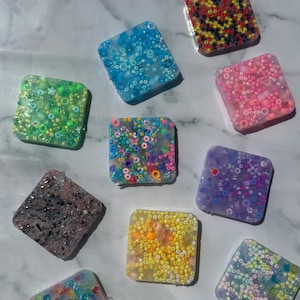 Könnte beinhalten: Eine Sammlung quadratischer Resin-Stücke, in die farbenfrohe Perlen eingebettet sind. Die Stücke variieren in der Farbe, darunter Grün, Blau, Rosa, Gelb und Lila. Die Perlen sind in verschiedenen Größen und Farbtönen gehalten und erzeugen ein strukturiertes, mosaikartiges Aussehen. Die Stücke werden auf einer hellen Oberfläche präsentiert.
