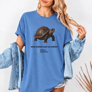 May include: A blue short-sleeved t-shirt featuring a brown tortoise graphic. Below the tortoise, the text reads "slow motion over no motion" followed by the National Geographic logo. The shirt has a crew neckline and a relaxed fit.
