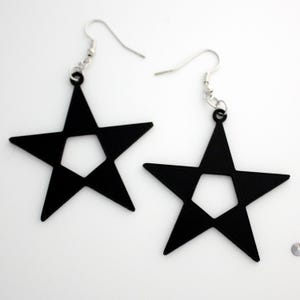 May include: A pair of black star-shaped earrings with silver-colored ear wires. Each earring features a five-pointed star design with a pentagon cut out in the center. The earrings are displayed against a white background.