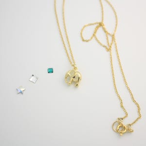 May include: A gold-toned necklace featuring a small elephant pendant. Beside the necklace are three loose decorative elements: a tiny square teal gemstone, a small clear square crystal, and a small iridescent star-shaped crystal piece.
