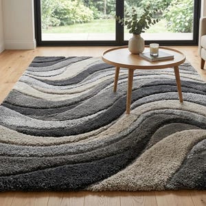 Puede incluir: Una alfombra rectangular con un patrón ondulado en tonos grises, beige y carbón. La alfombra está colocada sobre un suelo de madera, con una pequeña mesa redonda y un sofá en el fondo. El diseño de la alfombra añade un toque moderno a la habitación.
