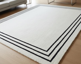 Può includere: Un tappeto rettangolare bianco con una superficie strutturata e un bordo nero. Il bordo è composto da tre linee nere parallele che incorniciano i bordi del tappeto. Il tappeto è posato su un pavimento in legno.
