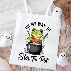 Puede incluir: Bolso de mano blanco con una rana de dibujos animados con un mono naranja removiendo un caldero. El texto "ON MY WAY TO Stir The Pot" está impreso en negro. Se ven dos pequeños peluches de fantasmas.