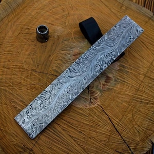 May include: A long, rectangular knife blade with a unique, swirling pattern in shades of gray and white. A small, cylindrical metal piece and a black leather strap are also visible. The blade rests on a wooden surface.